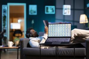 Man listens to music and reads from tablet, focus on stock market graphs on laptop screen. Teleworker wearing headphones, enjoying leisure time next to notebook showing trading investment charts photo