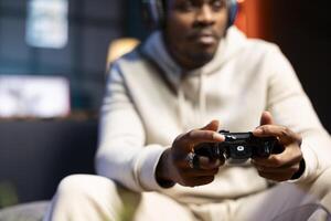 BIPOC man playing shooter videogame in neon lit apartment, holding controller, close up shot. African american gamer participating in internet multiplayer game using console system and headphones photo