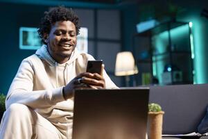 Upbeat man in apartment taking break from work to write messages on phone. Merry remote employee holding cellphone talking with mates over online texting app before continuing project on laptop photo