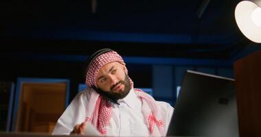 Muslim worker answering phone call while remotely working from cozy apartment office. Middle Eastern teleworker talking with colleague using smartphone, discussing paperwork and tasks needing to be solved photo