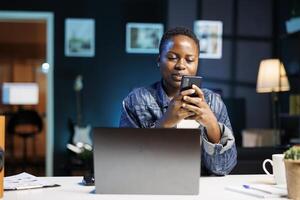 African american female blogger texting on mobile device while sitting in front of laptop on desk. Young black woman holding and using her smartphone, browsing the internet and social media. photo