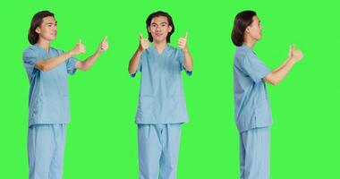 Healthcare nurse gives thumbs up sign against isolated greenscreen backdrop, showing like gesture and being pleased. Medical assistant presenting agreement and okay symbol. photo
