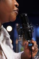 Black woman uses a professional microphone to record a podcast in home studio. Close-up of African individual touching and speaking into a modern mic while doing a radio broadcast. photo