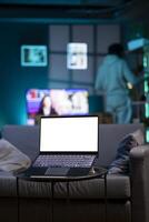 Focus on isolated screen laptop on coffee table in RGB lit home at night with man in blurry background looking on shelves. Close up shot of mockup notebook and person in the back doing inventory photo