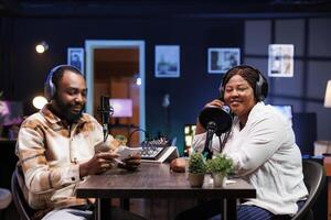 Black journalist uses modern recording technology to interview smiling female content creator in home studio. They broadcast their lively conversation to online audience via social media platforms. photo