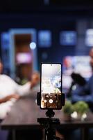 In a neon-lit studio, a vlogger show host is interviewing a guest while a smartphone on a tripod records them. The camera is focused on the phone capturing the content of the influencers. photo