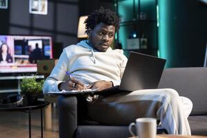 Self employed man working from home, writing business ideas, looking at laptop screen. BIPOC man sitting on couch with notebook in lap, using pen to note plan on paper while teleworking in apartment photo
