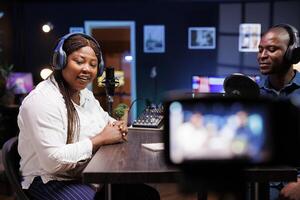 Talk show hosts in neon-lit home studio using professional equipment to create high quality content. Camera positioned on tripod films every move of african american couple doing a podcast episode. photo