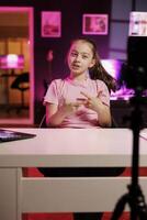 Cheerful child utilizes cellphone on tripod stabilizer to shoot introductory segment for vlog. Young content creator in pink neon studio welcomes devoted fan community, filming with mobile phone photo