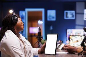 Black woman grasping device displaying blank white screen while hosting a talk show. Female content creator using headphones and podcast equipment while holding tablet with chromakey template. photo