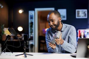 Smartphone on holder capturing footage of smiling reviewer discussing with viewers about V mount battery. Influencer chats with fans during stream, filming portable lithium accumulator photo