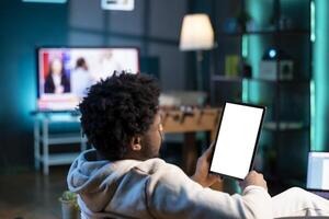 African american man sitting on couch, reading eBook from tablet in apartment using mockup tablet. Relaxed person enjoying interesting lecture on isolated screen device at home, relaxing during weekend photo