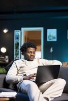 Smiling self employed man checking his emails on laptop while laying on couch. Cheerful remote employee working from home in bright stylish cozy apartment personal office, reading news on notebook photo