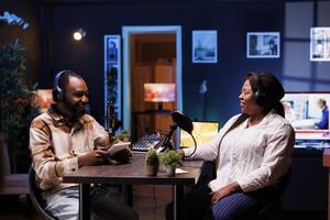 African podcasters having joyful discussion in home studio, exchanging various topics with online listeners. Show presenter with notebook is interviewing female socialite, having positive interactions photo