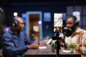 Selective focus of a smartphone on tripod being used to record two african american bloggers discussing different topics. Black men meeting in home studio using a mobile device to film a show. photo