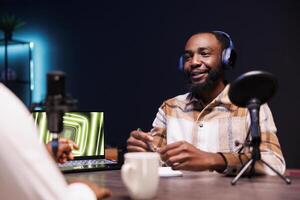 Black guy with a notebook is using podcast equipment to provide online advice. Male African American vlogger is seated at the table, recording an episode at home while speaking into a microphone. photo