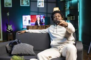 Man zapping through TV channels using remote, relaxing on couch in living room used as home theatre. BIPOC person sitting on sofa, using television set to entertain himself photo