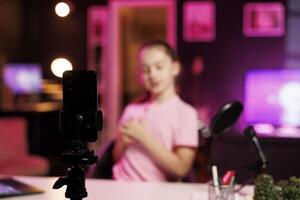 Focus on phone attached to tripod filming girl in home studio talking about day at school with children watching her. Kid in blurry background recording content for online channel with smartphone photo