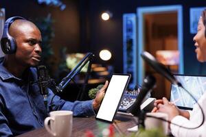 Black man holding device displaying blank white screen while speaking on a microphone. Male content creator using headset and podcast equipment while grasping tablet with copyspace mockup template. photo