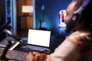 Selective focus on laptop displaying an isolated chromakey mockup template while black man uses podcast equipment. Podcaster makes live broadcast on streaming platform while device shows white screen. photo