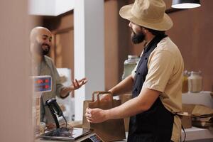 Middle eastern man interacting with caucasian vendor while waiting for his grocery package at checkout counter. Male vendor is packing organic bio food products in paper bag while customer waits. photo