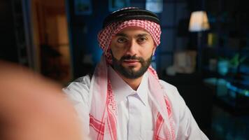 POV of cheerful Muslim content creator dressed in traditional clothing filming himself. Smiling Arab man with thobe and headscarf waving towards camera, doing online vlogging photo