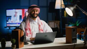 Portrait of happy Arab businessman working at home desk, typing on his laptop, solving tasks. Middle eastern man browsing on digital device, doing email communication in professional office setting photo