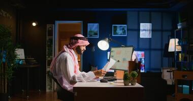 Muslim teleworker crosschecking analytics statistical data set with paperwork while remotely working in dimly lit apartment office. Hardworking Arab employee optimizing key performance indicators photo