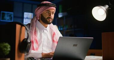 Arabic teleworker remotely working in dimly lit apartment sending emails on laptop. Middle Eastern worker at home typing business data on notebook device, wearing traditional attire photo