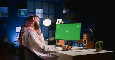 Arabic man looking at mockup computer screen while working from home. Self employed person typing on keyboard, writing emails on isolated screen device in apartment office with TV as background noise photo