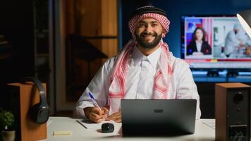 Portrait of smiling Arab entrepreneur working at home office desk on laptop, browsing the internet and writing in notebook. Middle Eastern man using pen, notepad and digital device while teleworking photo