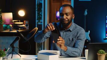 African american tech content creator filming technology review of newly released smartphone, unboxing it and presenting specifications to audience. blogger showing phone to subscribers photo