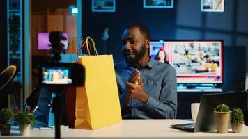 Content creator holding shopping bags, filming haul in dimly lit studio using vlog recording devices. Internet star doing of clothing items he bought using professional camera photo