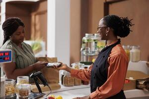 African American woman happily makes cashless payment at market counter using credit card. She smiles as she communicates with the vendor and completes the transaction with a debit and pos machine. photo