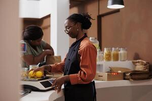 An aproned African American woman assists a customer at a fresh food market with measuring the weight of lemons on digital scale. Female seller weighing locally grown bio produce. photo