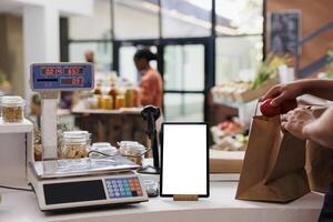 Digital tablet with a white screen display is placed vertically on counter next to a weighing scale. Electronic device showing isolated mockup template while the vendor packs food products in a bag. photo