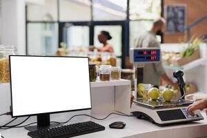 Desktop computer showing blank white screen while a salesperson measures apples using a weighing scale. Positioned at cashier desk is a pc monitor displaying isolated chromakey mockup template. photo
