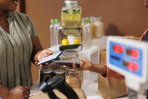 Closeup of african american client using mobile device on pos machine to pay for fresh organic product at local grocery store. Black woman making nfc payment with cell phone for bio food items. photo