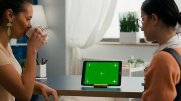 Two collegues talking about internet connection in front of tablet computer with mock up green screen chroma key. Women browsing on isolated device sittings at office desk in modern living room photo