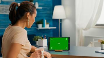 Caucasian female looking at tablet computer with mock up green screen chroma key sitting on office desk in living room. Freelancer woman browsing on social network using isolated device photo