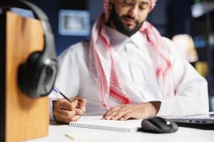 Selective focus on an Arab student writing on his notebook with his laptop nearby. Close-up shot of a Middle Eastern guy using a pen to jot down his notes from his personal computer. photo