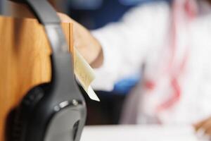 Selective focus of a sticky note and wireless headphones placed on a wooden stand by a Middle Eastern person. Close-up shot of Arab man putting a written paper on a board as a reminder. photo