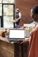 Female customer browsing on laptop with an isolated white screen. African american woman using minicomputer displaying blank chromakey mockup template in an eco friendly sustainable store. photo