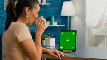 Freelancer woman holding cup of coffee looking at tablet computer with mock up green screen chroma key sitting on desk table. Caucasian female browsing on isolated device from home office room photo