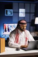 Middle Eastern man in traditional attire working indoors, using a laptop for browsing while engaged in call, using wireless headphones. Image shows male Arab person utilizing modern technology. photo