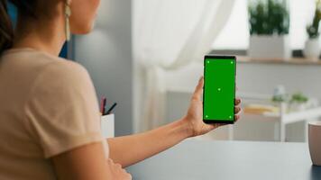 Back view of freelencer holding chroma key green screen mock up smartphone watching social media content. Caucasian woman sitting on office desk working on marketing project using isolated gadget photo