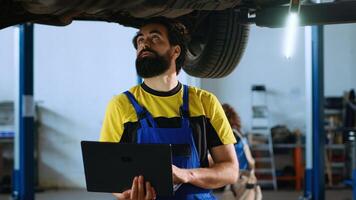 Licensed mechanic standing underneath suspended car in garage workspace using laptop to order new parts after finding issues. Employee looking online for replacing components using device photo