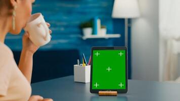 Woman drinking a cup of coffee while looking at tablet computer with mock up green screen chroma key sitting on office table. Freelancer using with isolated gadget for internet communication project photo