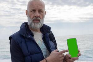 Mockup image of a grey-haired bearded man pointing his finger at a mobile phone with a blank green screen. High quality photo