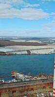 Survey of heat power station from aerial view on the background of long river and private sector with houses within the city. Environmental pollution. Vertical video